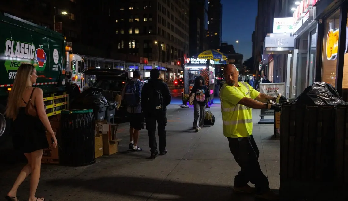 A Classic Recycling driver wheeling a container to the truck on a Manhattan sidewalk at night